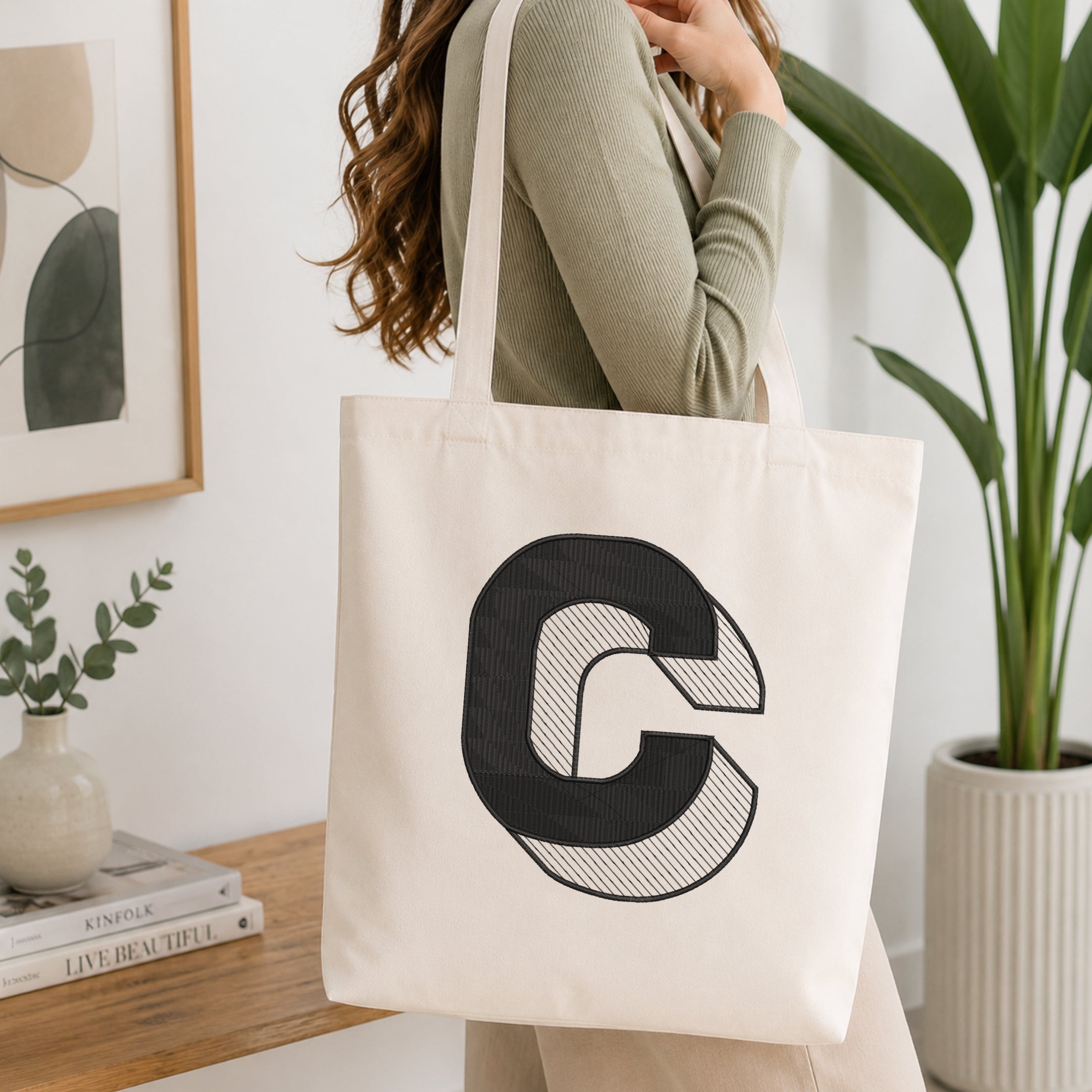 A woman wearing a sage green ribbed long-sleeve top carries a cream canvas tote bag with a large black stylized letter 'C' printed on the front, set in a bright, modern room with a wooden table, framed art, and a potted plant.