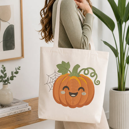 A person wearing a sage green ribbed long-sleeve top carries a cream canvas tote bag featuring a cute orange pumpkin with a smiling face, green stem, and spiderweb graphic, set against a bright, modern room background with a wooden table and potted plants.