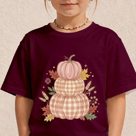 A child wearing a dark burgundy t-shirt with a graphic of three stacked pumpkins in plaid patterns, surrounded by autumn leaves and stars.