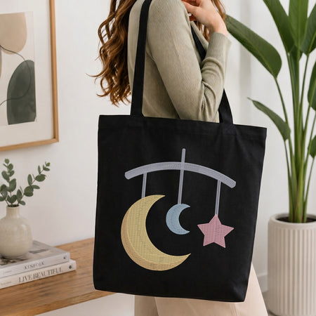 A woman wearing a beige ribbed long-sleeve top carries a black canvas tote bag with a celestial mobile design featuring a yellow crescent moon, a light blue crescent moon, and a pink star.