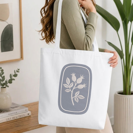  Woman carrying a white natural canvas tote bag featuring an embroidered botanical rosehip design in a bright, modern interior setting.   