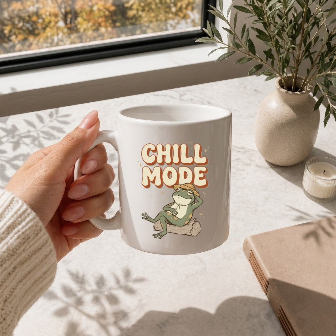 Hand holding a mug with 'Chill Mode' text and frog graphic on a table with a window and plant in the background.