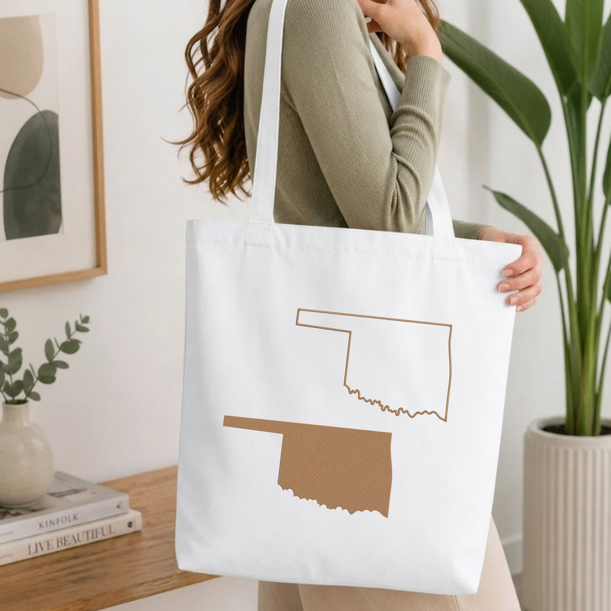 Woman carrying a white natural canvas tote bag featuring an embroidered Oklahoma state map design in a bright, modern interior setting.
