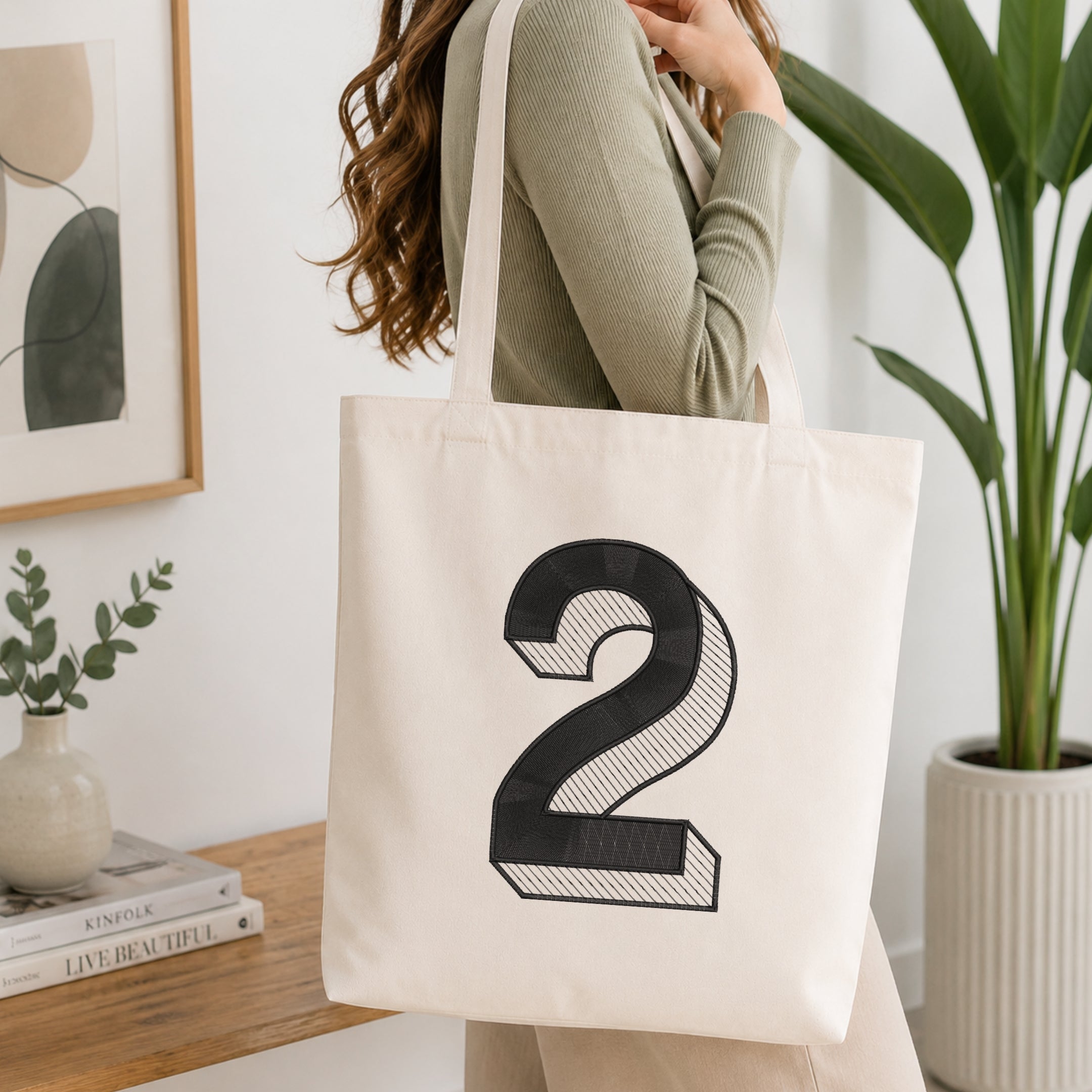 A woman wearing a sage green ribbed long-sleeve top carries a cream canvas tote bag with a large black number 2 printed on the front, set in a bright, modern room with a wooden table, framed art, and a potted plant.