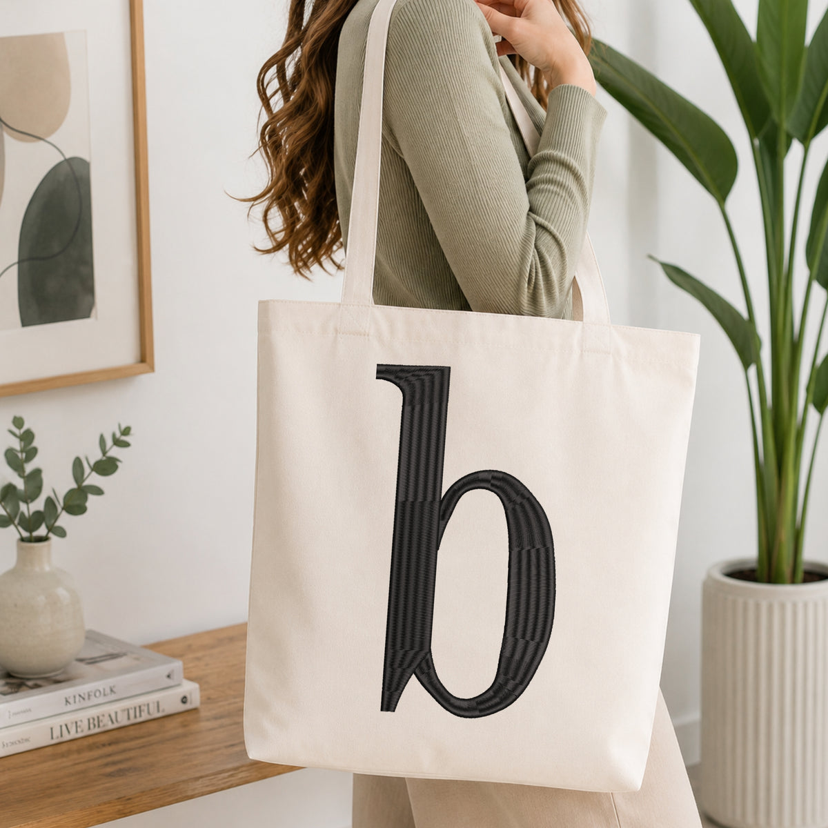 A woman wearing a sage green ribbed long-sleeve top carries a cream canvas tote bag with a large black embroidered letter 'b' on the front, set in a bright, modern room with a wooden table, books, and a potted plant.