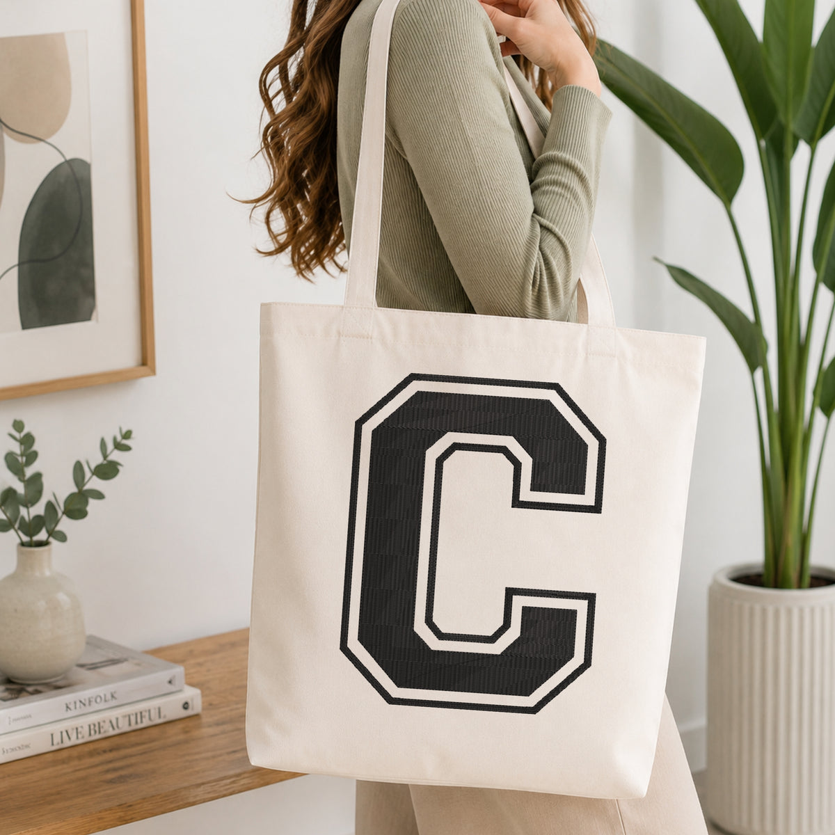 A woman wearing a sage green ribbed long-sleeve top carries a cream canvas tote bag with a large black collegiate letter 'C' outlined in white on the front, set in a bright, modern room with a wooden console table, framed art, and a potted plant.