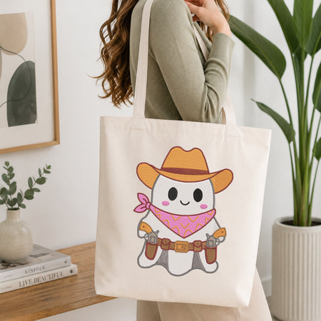 A woman wearing a sage green ribbed sweater carries a cream canvas tote bag with a cute cartoon ghost cowboy graphic. The graphic features an orange cowboy hat, pink bandana, and a gray body.