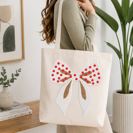 A woman wearing a sage green ribbed sweater carries a cream canvas tote bag decorated with a large white and red bow design featuring brown ribbon details, set against a bright, modern interior background with a wooden table and potted plants.