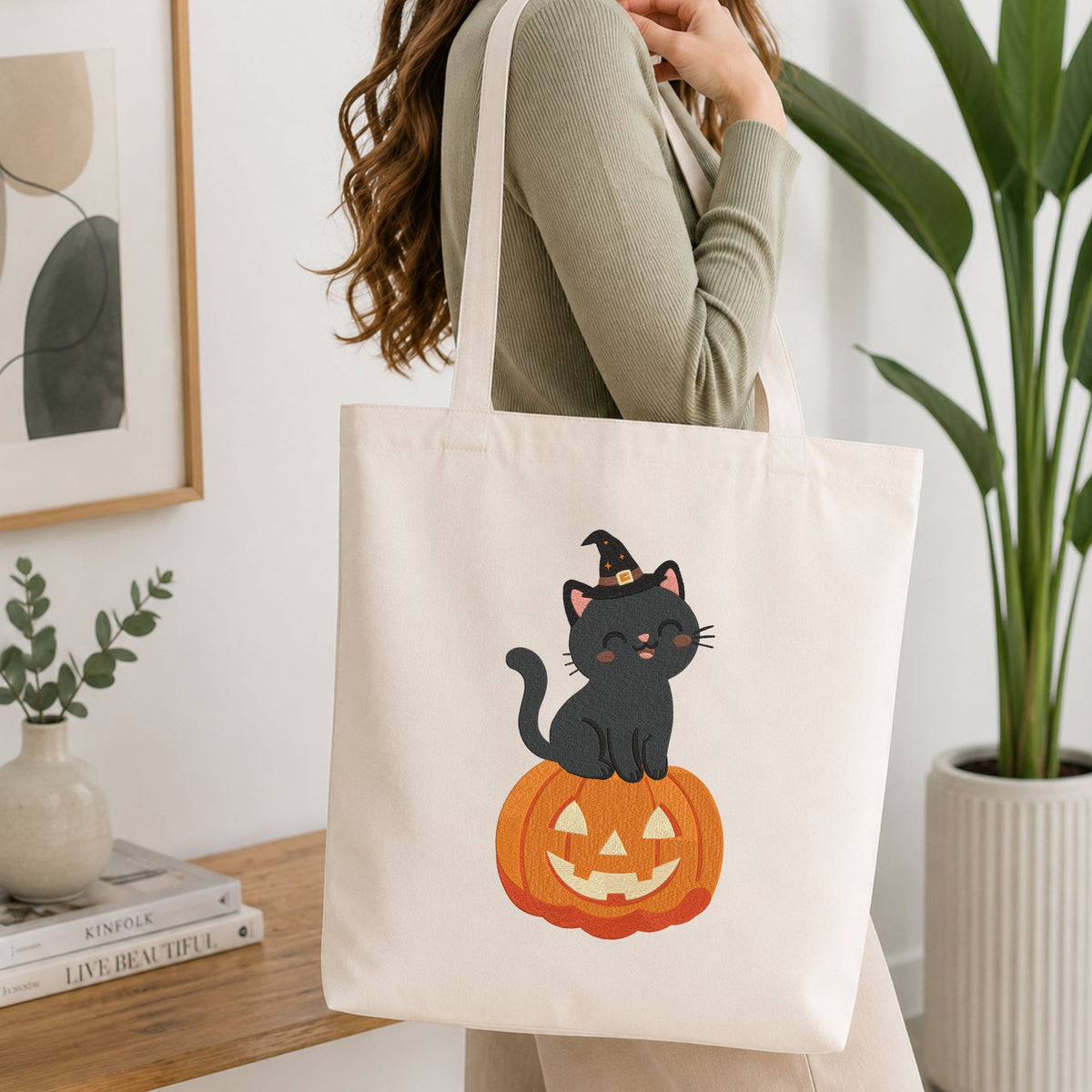 Woman carrying a white natural canvas tote bag featuring a cute black cat wearing a witch hat sitting on a jack-o-lantern pumpkin machine embroidery design in a bright, modern interior setting