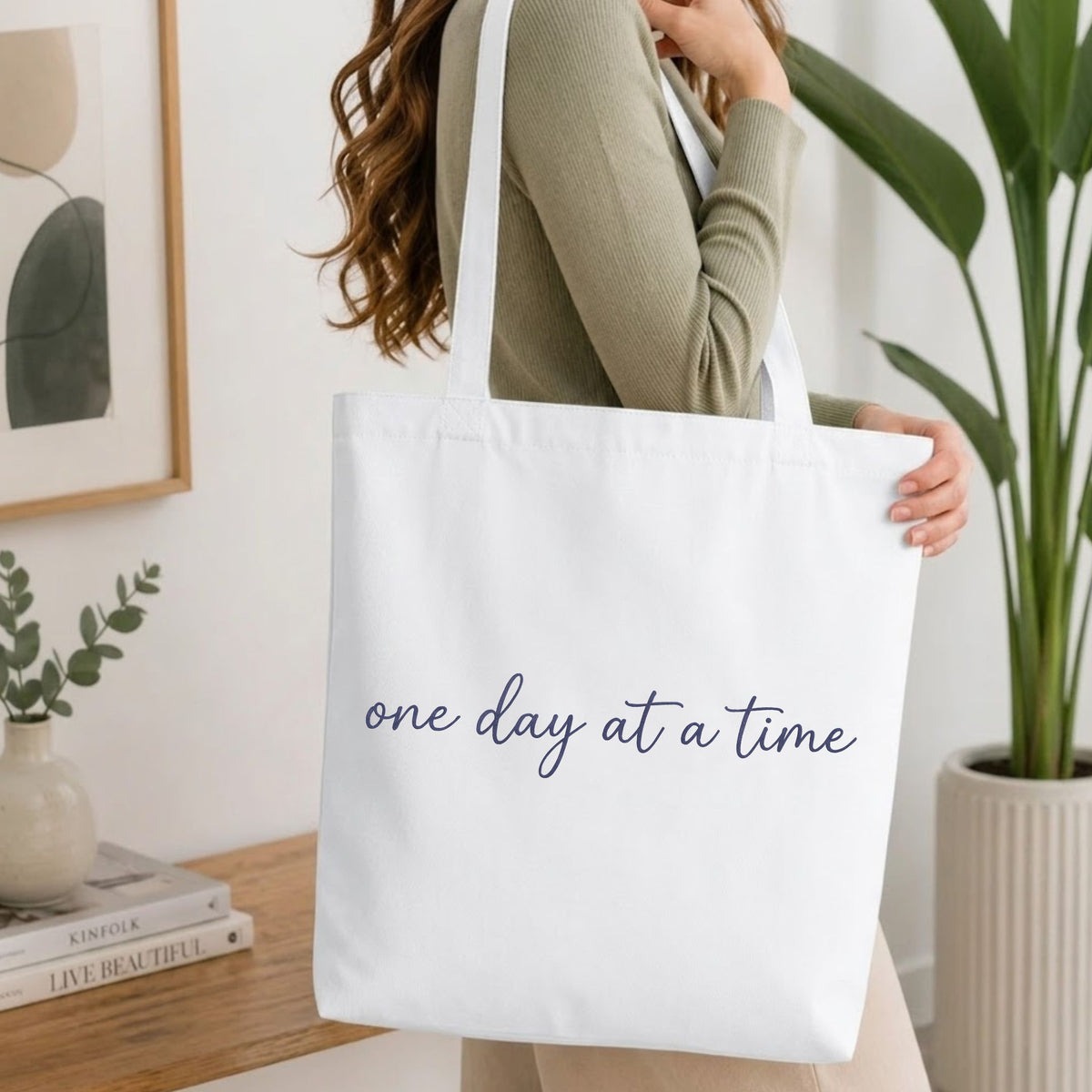 A woman wearing a sage green ribbed sweater carries a white canvas tote bag with a large tan bone outline graphic on the front, set in a bright, modern room with a wooden table and potted plants.