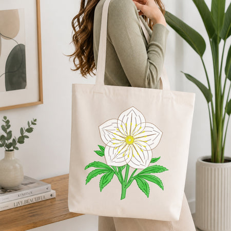 A woman wearing a sage green ribbed sweater carries a cream canvas tote bag with a large embroidered white flower with green leaves and a yellow center, set against a bright, modern interior background with a wooden table and potted plants.
