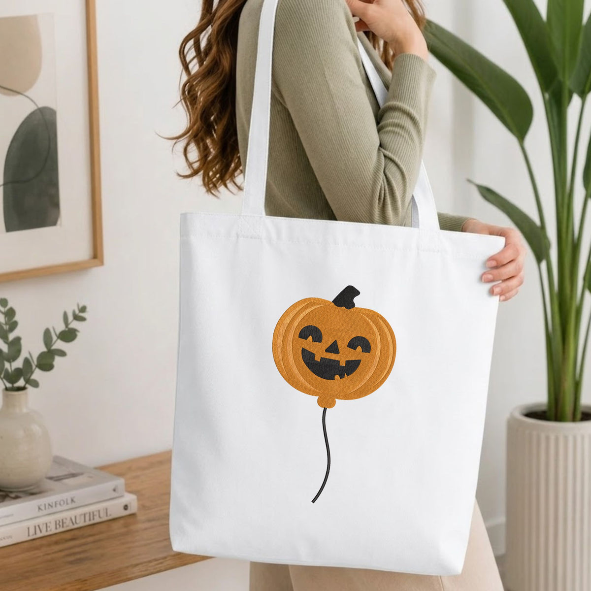 Woman carrying a white natural canvas tote bag featuring a cute ghost machine embroidery design in a bright, modern interior setting   