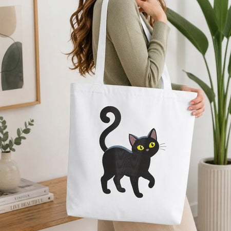 A woman wearing a sage green ribbed long-sleeve top carries a white canvas tote bag featuring a cartoon illustration of a black cat with yellow eyes, set against a bright, modern indoor background with a wooden table and potted plants.