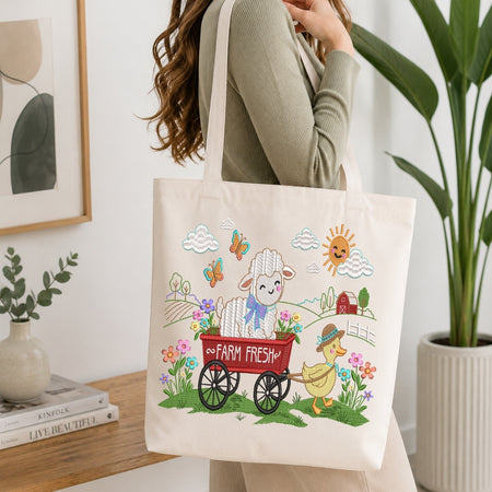 A woman carries a cream canvas tote bag with a colorful cartoon farm illustration featuring a white lamb in a red wagon, a yellow duck, flowers, butterflies, clouds, and a smiling sun. The scene is set in a bright, modern room with a wooden table, books, a framed print, and a potted plant.