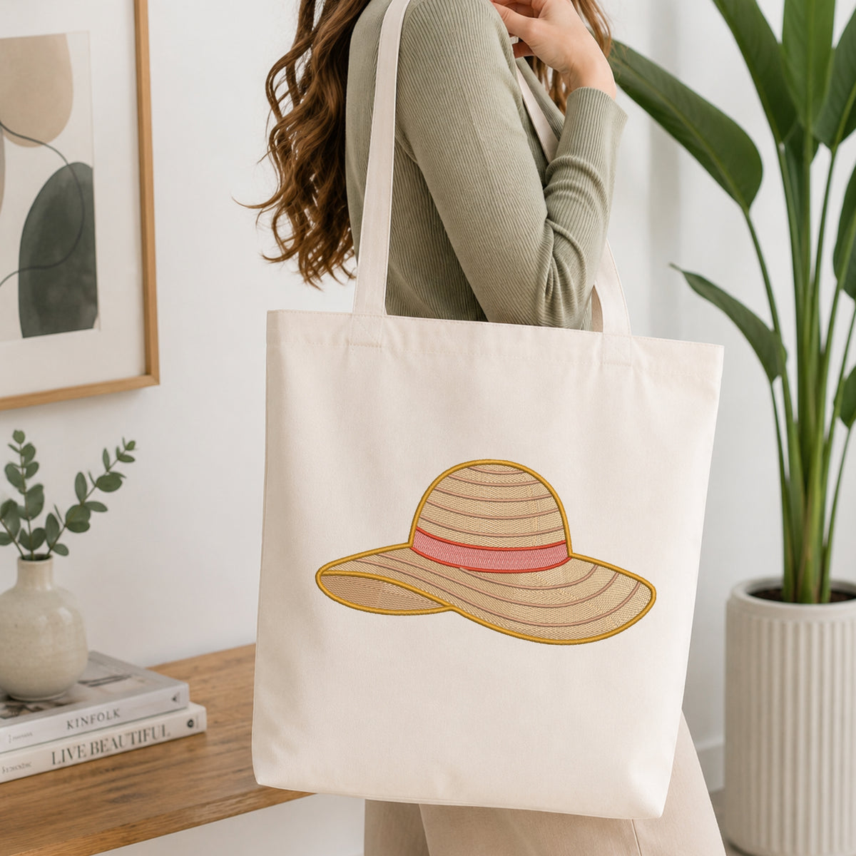 A woman wearing a sage green ribbed top carries a cream canvas tote bag with an embroidered straw hat design featuring a pink band, in a bright, modern room with a wooden table and potted plants.
