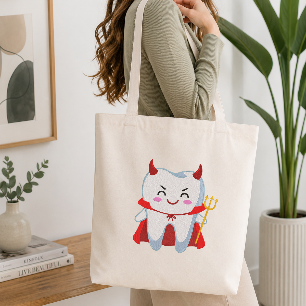 Woman carrying a white natural canvas tote bag featuring a cute devil tooth machine embroidery design in a bright, modern interior setting   