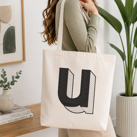 A woman wearing a sage green ribbed long-sleeve top carries a large off-white canvas tote bag with a bold black letter 'U' graphic on the front, set in a bright, modern room with a wooden table, framed art, and a potted plant.