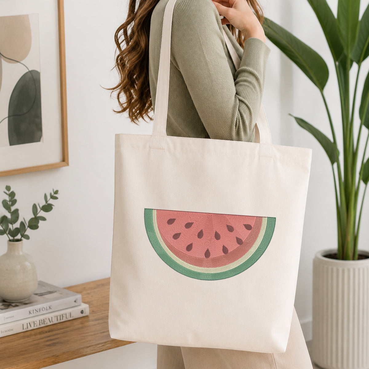 A woman wearing a sage green ribbed long-sleeve top carries a cream canvas tote bag featuring a watermelon slice graphic. The setting is a bright, modern room with a wooden console table, a potted green plant, and a framed abstract print.