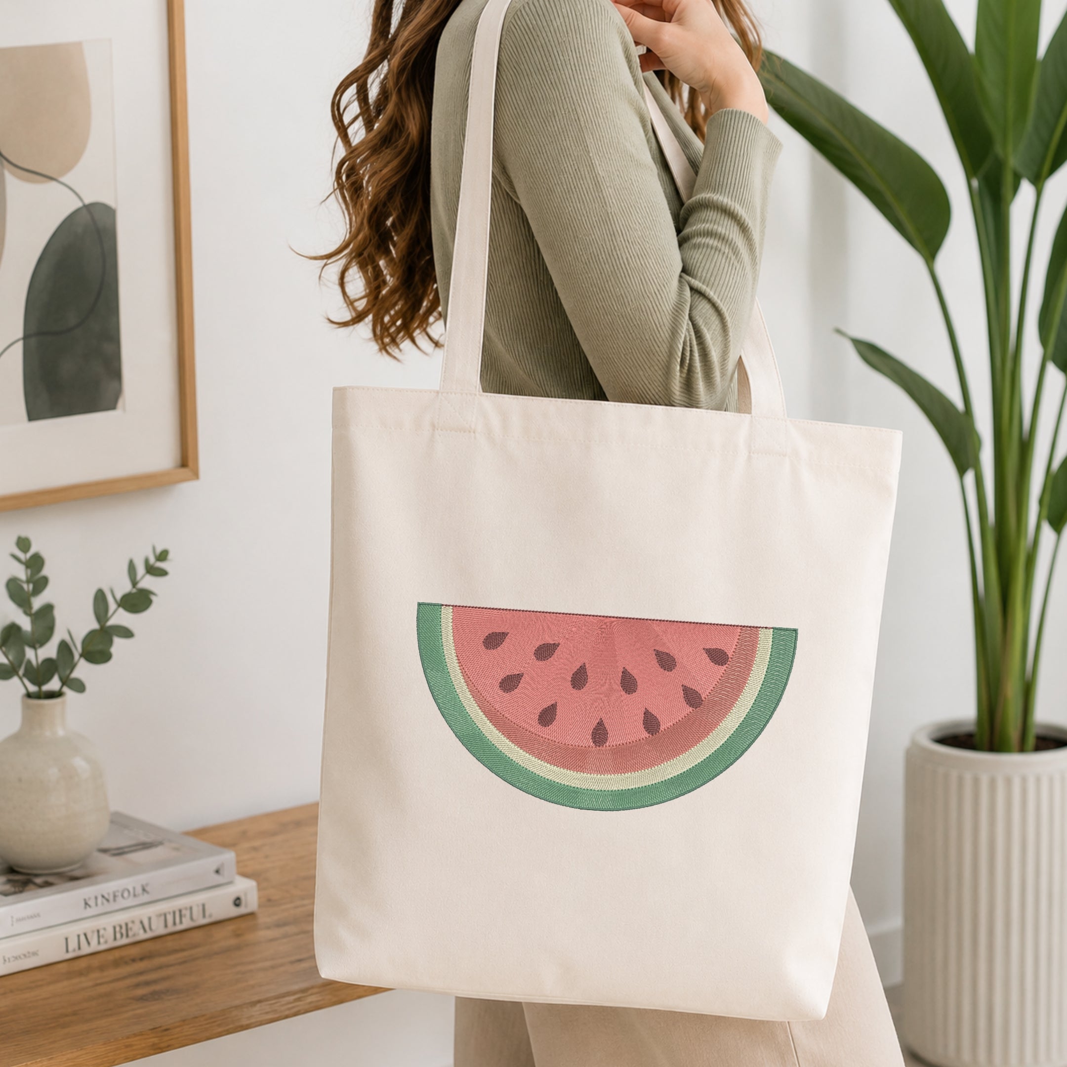 A woman wearing a sage green ribbed long-sleeve top carries a cream canvas tote bag featuring a watermelon slice graphic. The setting is a bright, modern room with a wooden console table, a potted green plant, and a framed abstract print.
