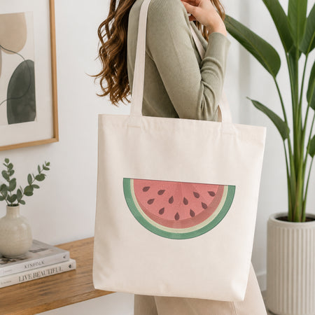 A woman wearing a sage green ribbed long-sleeve top carries a cream canvas tote bag featuring a watermelon slice graphic. The setting is a bright, modern room with a wooden console table, a potted green plant, and a framed abstract print.
