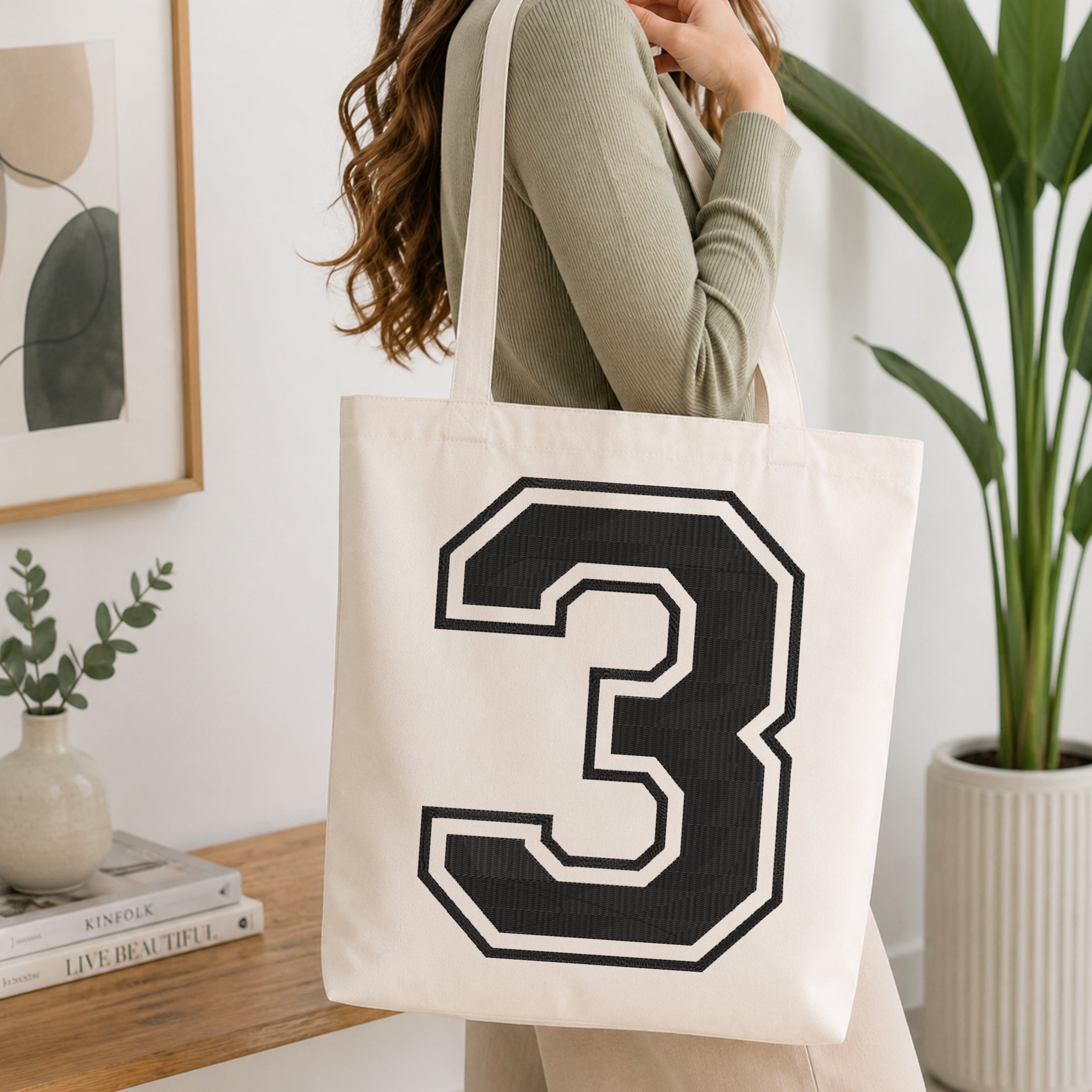 A person wearing a sage green ribbed long-sleeve top carries a cream canvas tote bag with a large black and white number 3 on the front, set in a bright, modern room with a wooden console table and a potted plant.