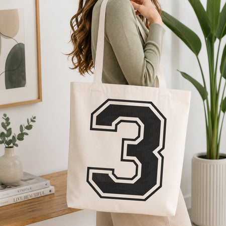 A person wearing a sage green ribbed long-sleeve top carries a cream canvas tote bag with a large black and white number 3 on the front, set in a bright, modern room with a wooden console table and a potted plant.