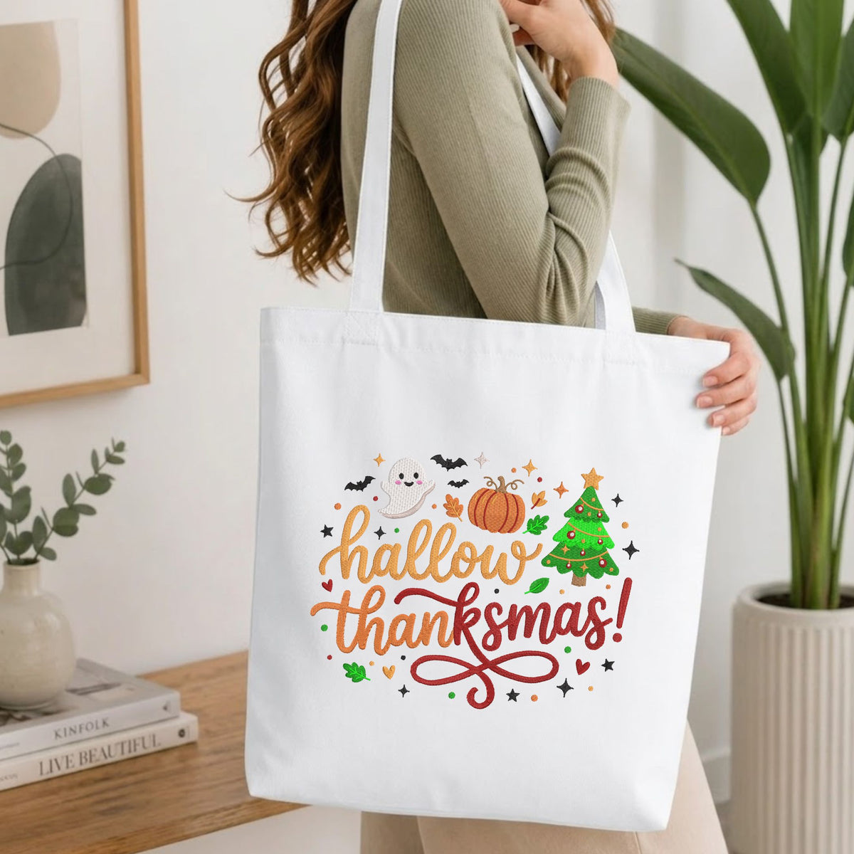  Woman carrying a white natural canvas tote bag featuring an embroidered "Hallowthanksmas!" holiday design in a bright, modern interior setting   