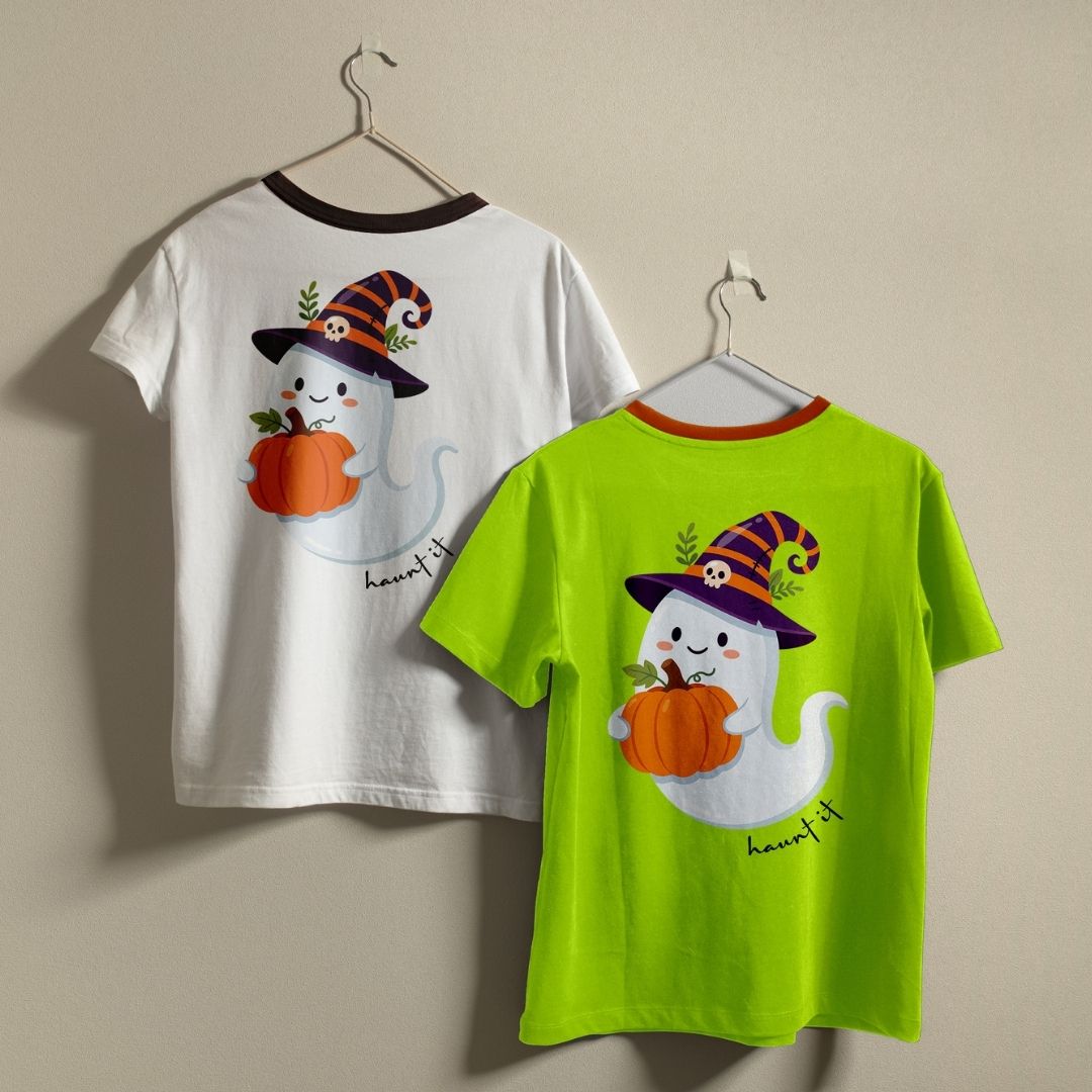 Two children's short-sleeve T-shirts hanging on hangers against a plain beige wall. The left shirt is white with a dark brown collar and features a ghost holding an orange pumpkin wearing a witch hat graphic on the front.