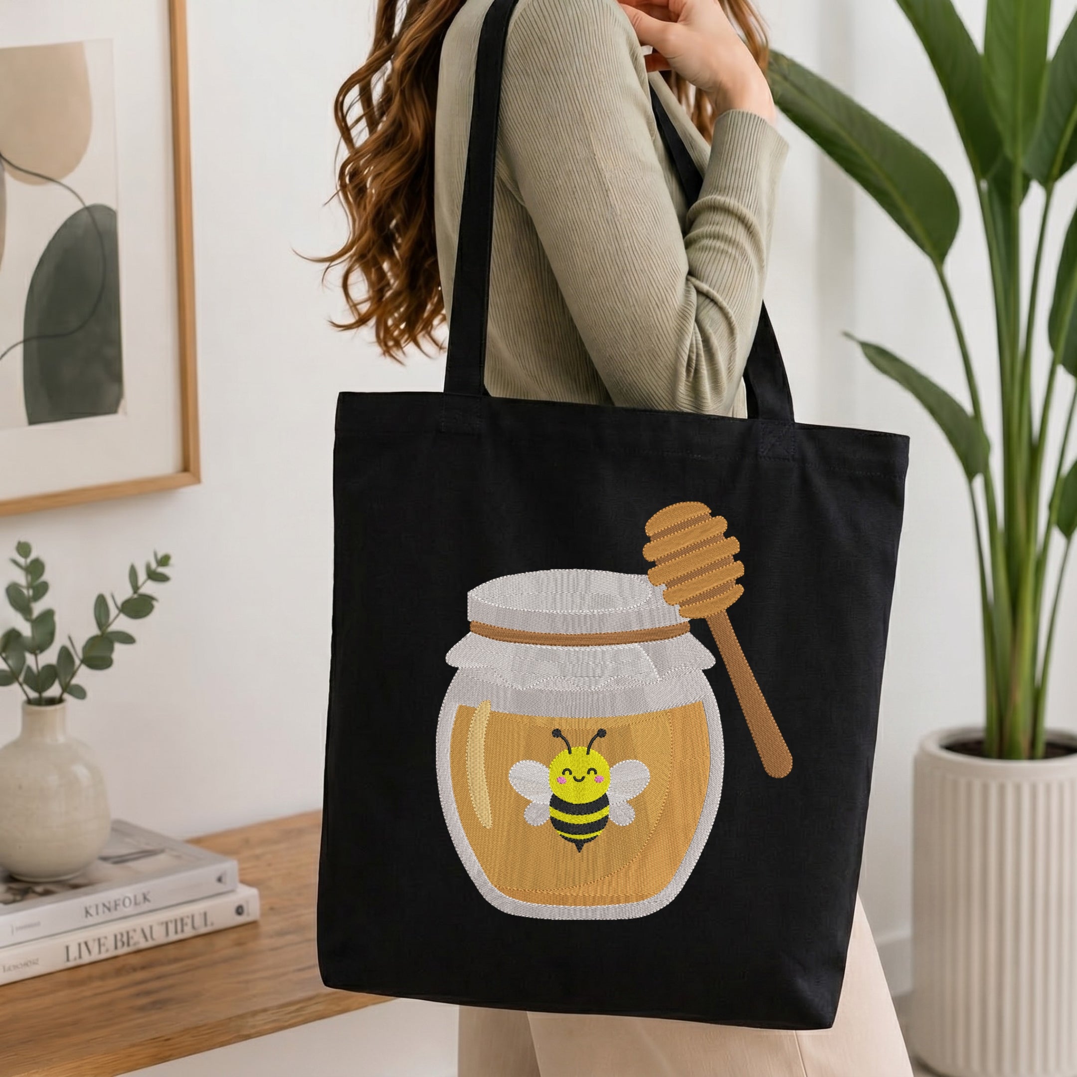 A woman wearing a beige ribbed sweater carries a black canvas tote bag with a printed honey jar and bee design. The scene is set in a bright, modern room with a wooden table, books, a small plant, and a potted green plant in the background.
