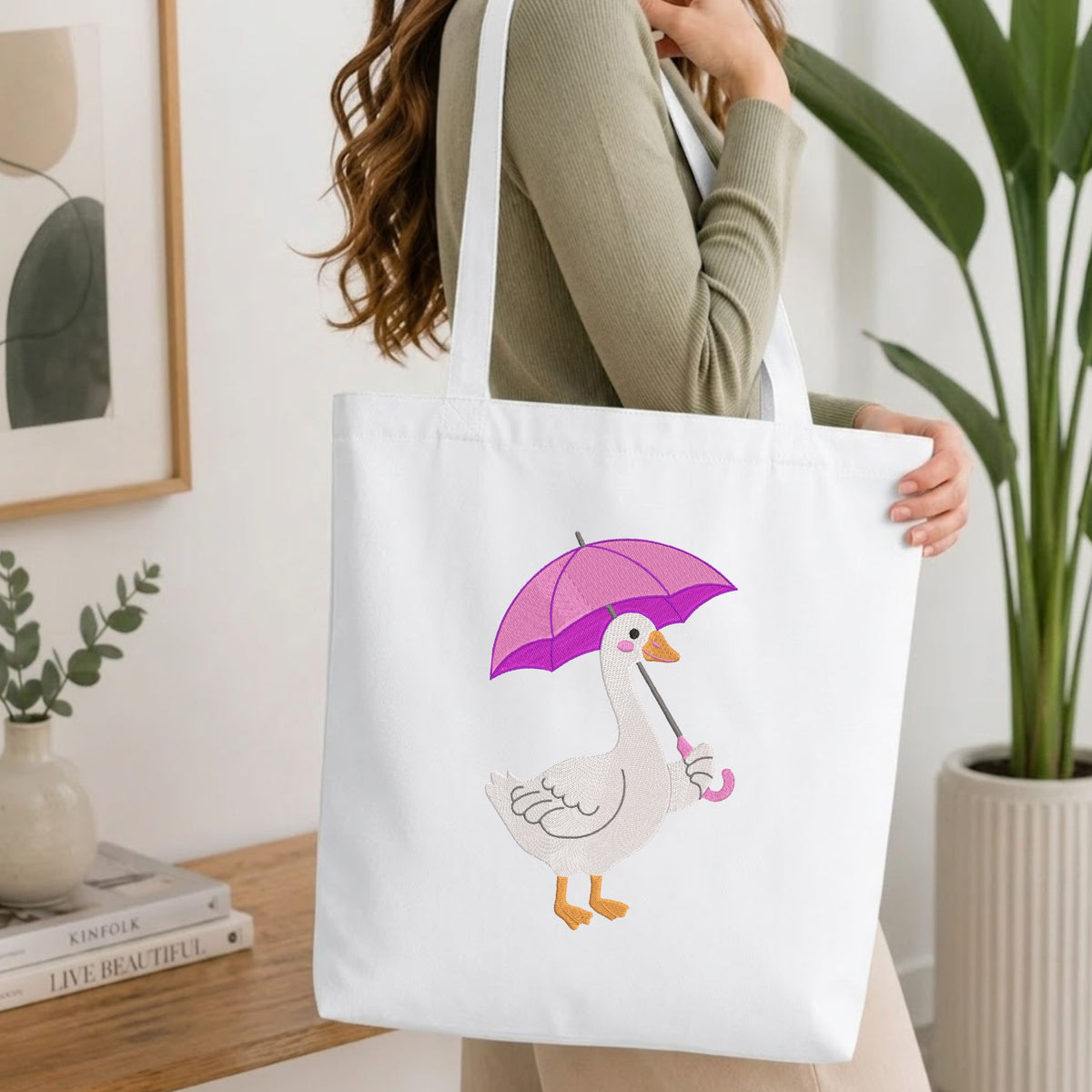 A woman wearing a sage green ribbed sweater carries a white canvas tote bag. The bag features a cute cartoon illustration of a white goose holding a pink and purple umbrella.