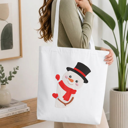 A woman wearing a sage green ribbed sweater carries a white canvas tote bag with a cartoon snowman design. The snowman has a black top hat with a red band, a red scarf, and a carrot nose.