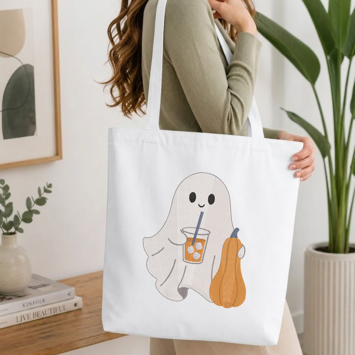 Woman carrying a white natural canvas tote bag featuring an embroidered cute ghost with iced coffee and pumpkin design in a bright, modern interior setting.   