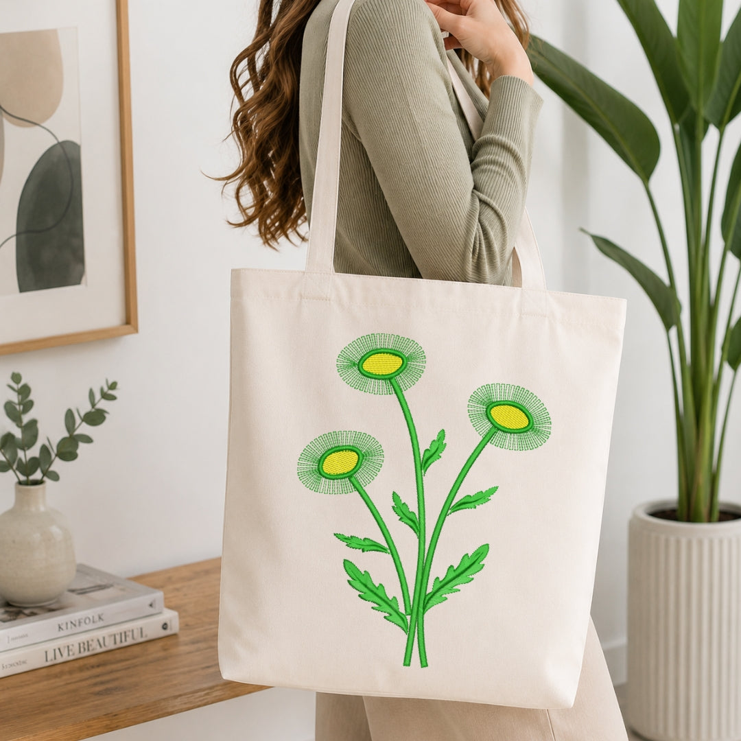 A woman wearing a sage green ribbed sweater carries a cream canvas tote bag with a green and yellow dandelion design, set in a bright, modern room with indoor plants and a wooden console table.