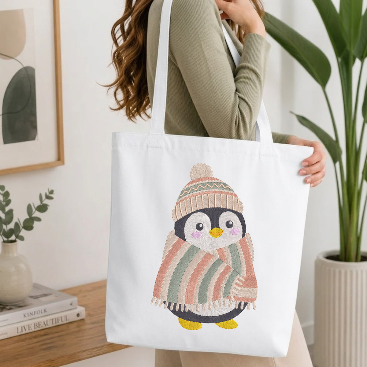 A woman wearing a sage green ribbed sweater carries a white canvas tote bag with a cute penguin illustration. The penguin is wrapped in a striped pink, orange, and green blanket and wears a knit hat.