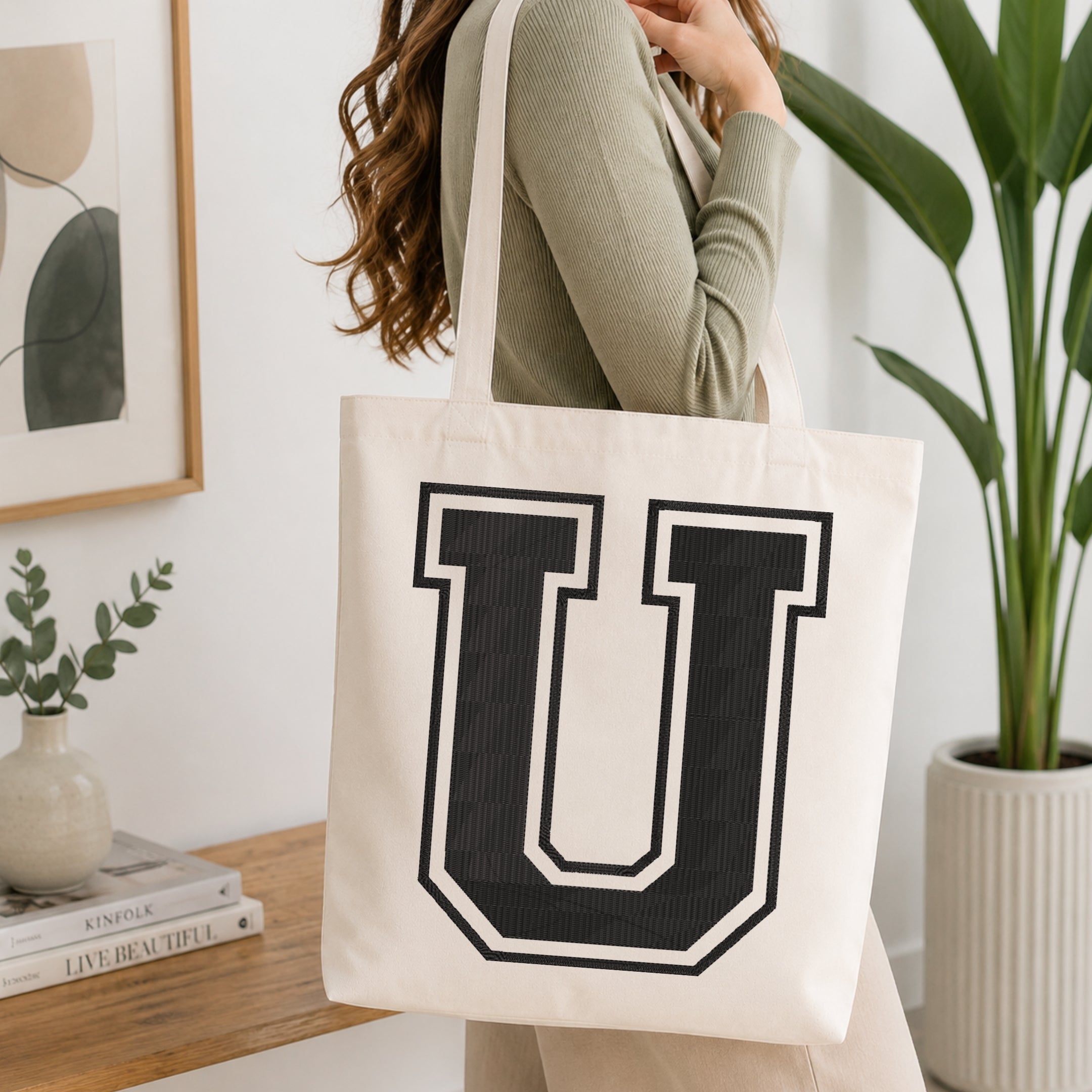 A woman wearing a sage green ribbed long-sleeve top carries a large off-white canvas tote bag with a bold black collegiate letter 'U' printed on the front, set in a bright, modern room with a wooden console table and a potted plant.