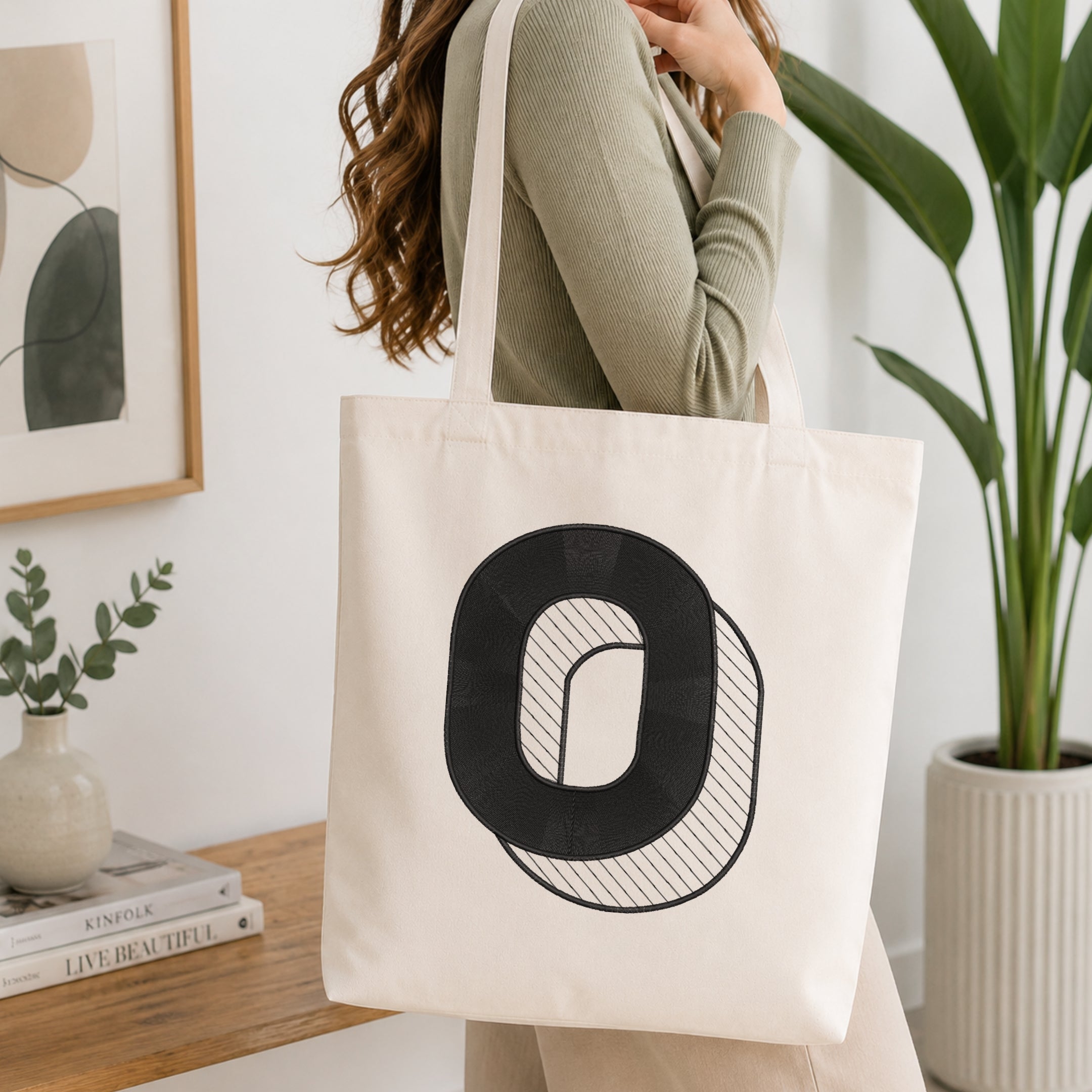 A woman wearing a sage green ribbed top carries a cream canvas tote bag with a large black letter 'O' graphic on the front, set in a bright, modern room with a wooden table, framed art, and a potted plant.