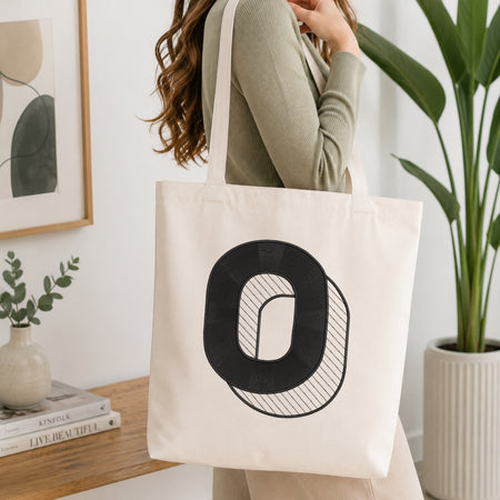 A woman wearing a sage green ribbed top carries a cream canvas tote bag with a large black letter 'O' graphic on the front, set in a bright, modern room with a wooden table, framed art, and a potted plant.