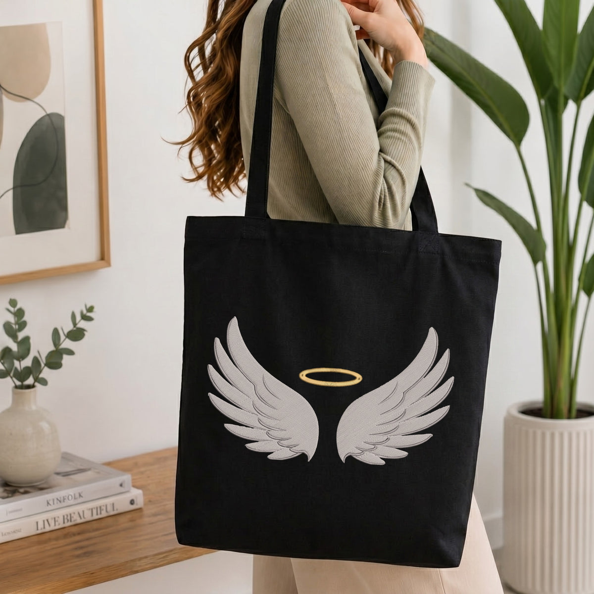 A woman wearing a beige ribbed sweater carries a black canvas tote bag with a white angel wings and halo design on the front, set in a bright, modern room with a wooden table and potted plants.