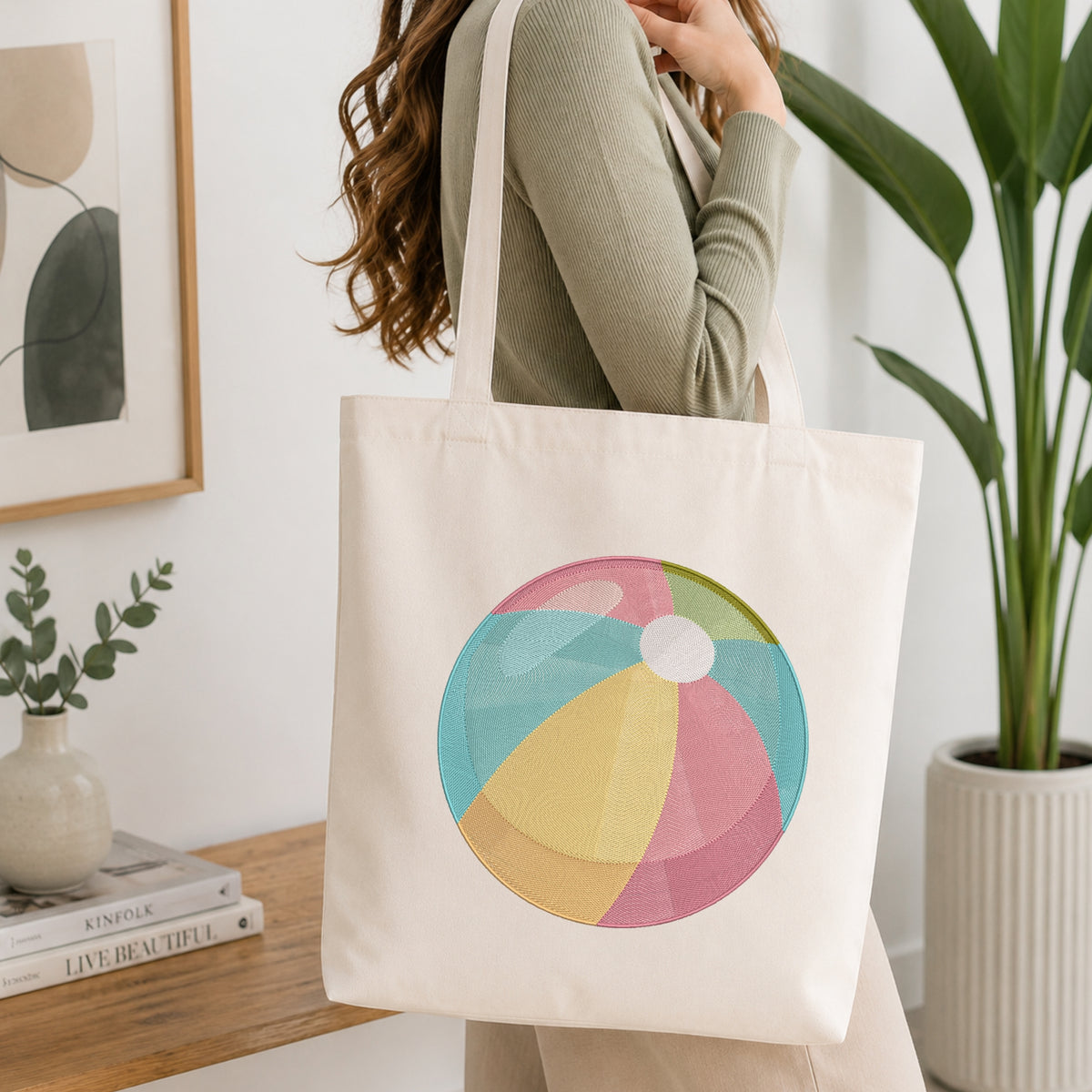A woman wearing a sage green ribbed long-sleeve top carries a cream canvas tote bag with a colorful beach ball graphic on the front, set in a bright, modern room with a wooden table, books, and a potted plant.