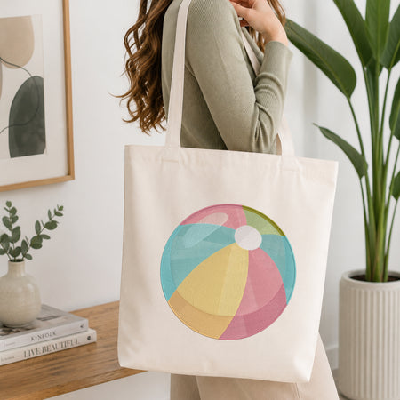 A woman wearing a sage green ribbed long-sleeve top carries a cream canvas tote bag with a colorful beach ball graphic on the front, set in a bright, modern room with a wooden table, books, and a potted plant.