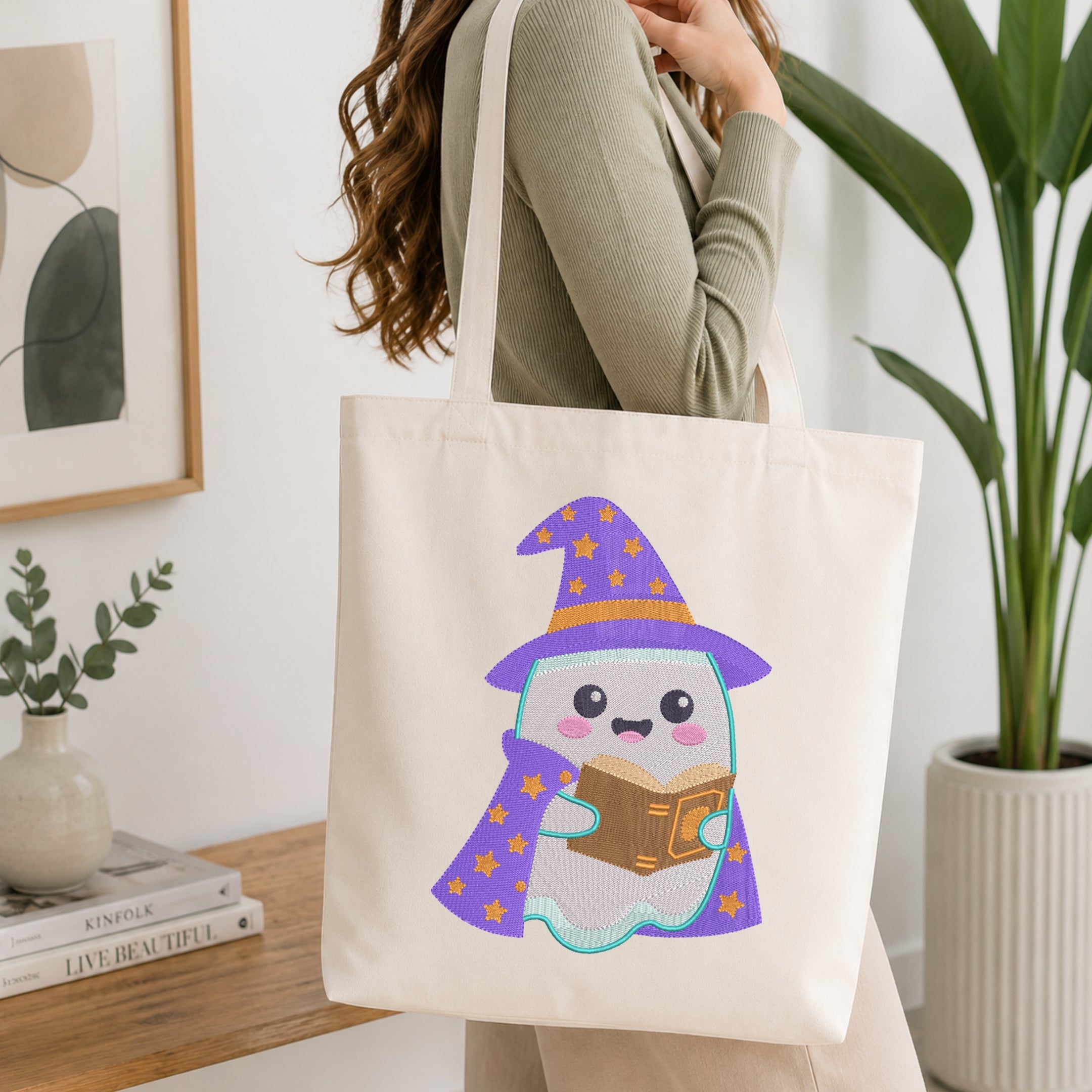 A woman holds a cream canvas tote bag with a cute cartoon ghost wizard graphic wearing a purple hat and cape. The scene is set in a bright, modern room with a wooden table, books, a small plant, and a large green potted plant in the background.