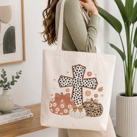 A woman wearing a sage green ribbed sweater carries a cream canvas tote bag with a fall harvest design. The design features a cross with black-and-white polka dots, two pumpkins with leopard print and white daisy patterns, and small autumn leaves.