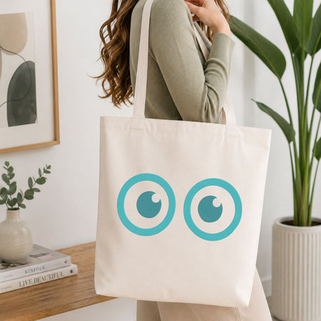 A woman wearing a sage green ribbed long-sleeve top carries a cream canvas tote bag with a graphic of two turquoise and white cartoon eyes. The setting is a bright, modern room with a wooden table, books, a framed print, and a potted plant.