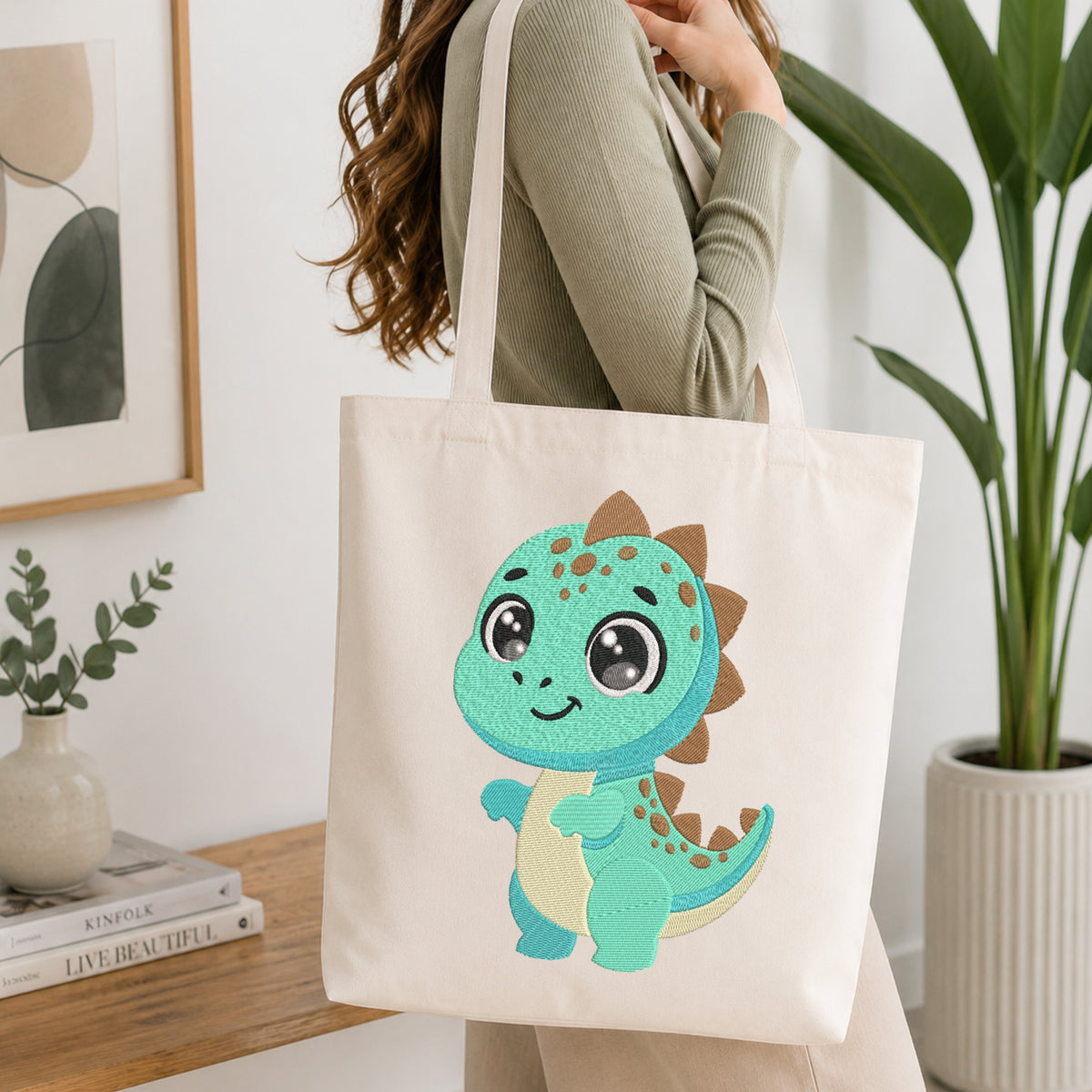 A woman wearing a sage green ribbed sweater carries a cream canvas tote bag featuring a cute turquoise and cream cartoon dinosaur with brown spikes. The scene is set in a bright, modern room with a wooden table, books, a framed print, and a potted plant.
