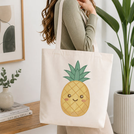 A person wearing a sage green ribbed long-sleeve top carries a cream canvas tote bag featuring a cute embroidered pineapple design with green leaves and a smiling face, set in a bright, modern room with a wooden table and potted plants.