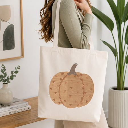 A woman wearing a sage green ribbed sweater carries a cream canvas tote bag with a large wooden pumpkin design featuring small hearts. The scene is set in a bright, modern room with a wooden table, books, a framed print, and a potted plant.