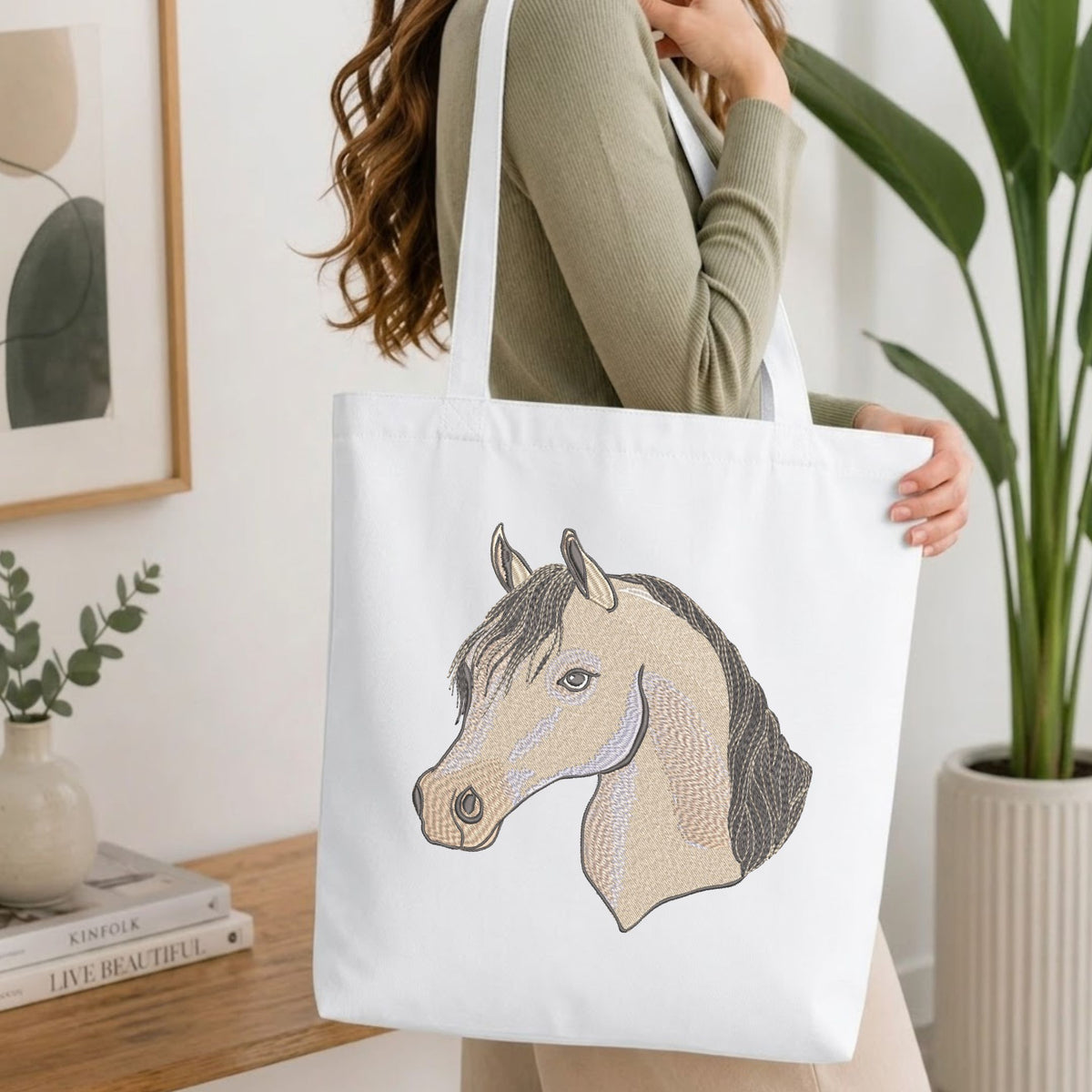 Woman carrying a white natural canvas tote bag featuring an embroidered elegant horse head design in a bright, modern interior setting