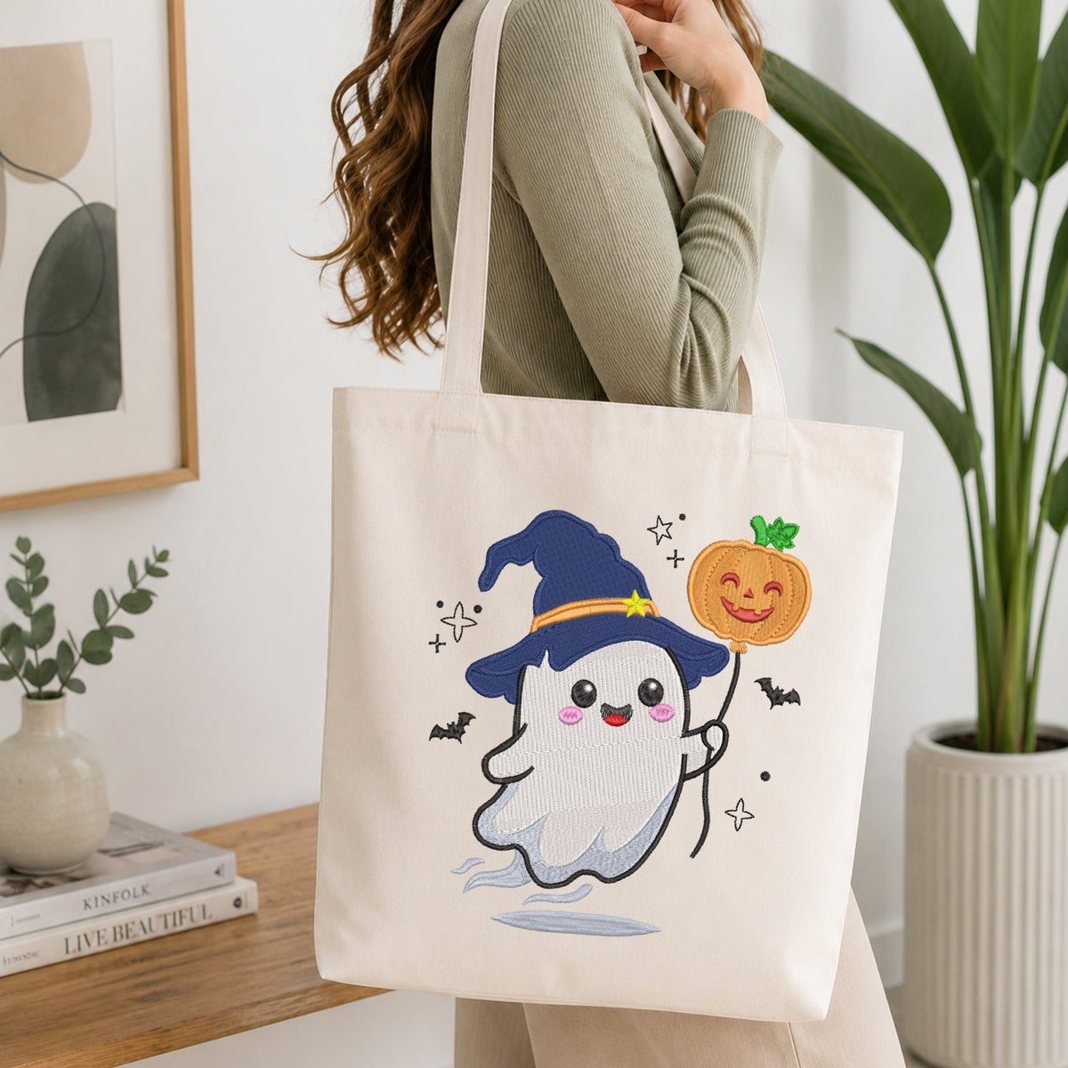 A woman wearing a sage green long-sleeve top carries a cream canvas tote bag decorated with a cute cartoon ghost wearing a blue witch hat and holding an orange pumpkin.