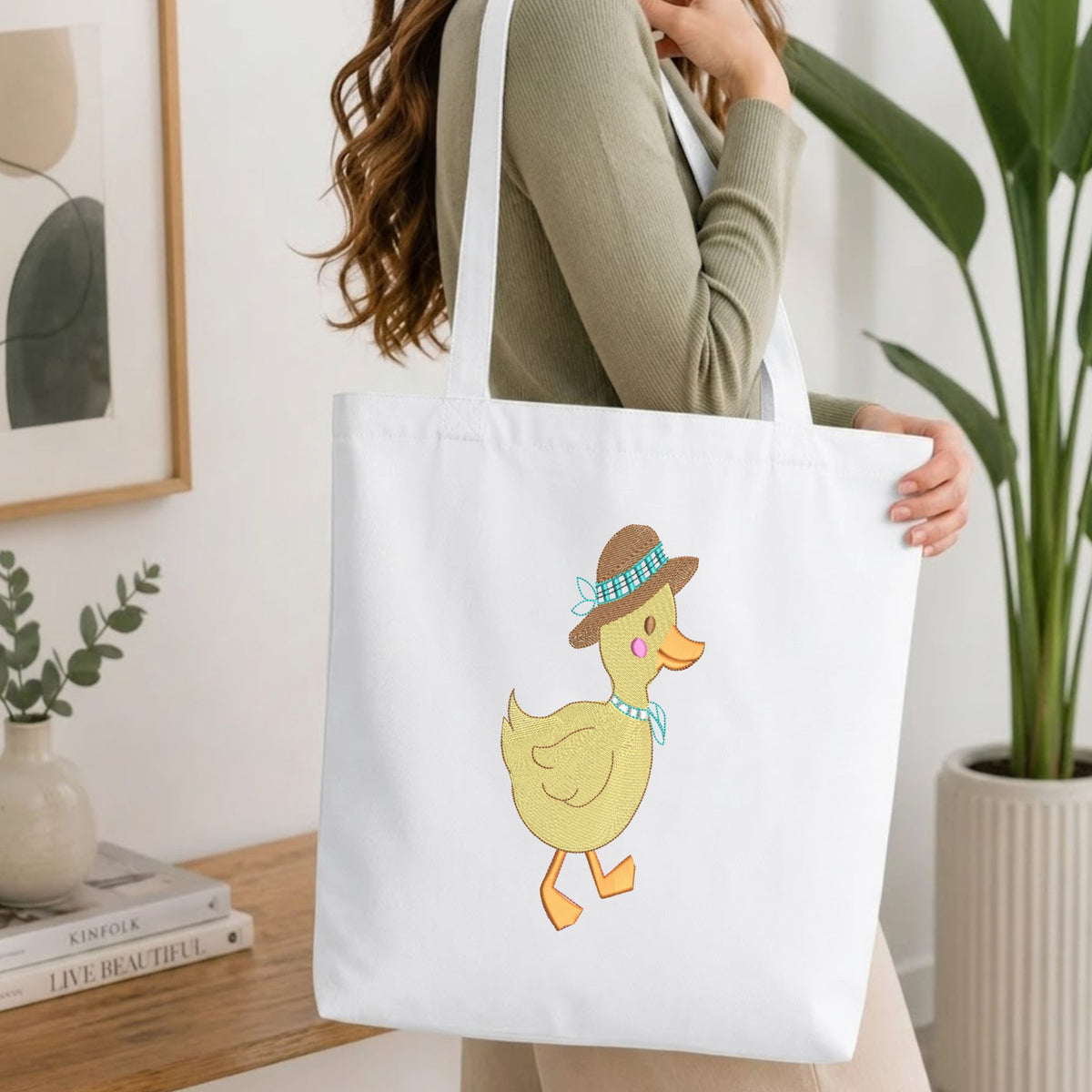 A woman wearing a sage green ribbed top carries a white canvas tote bag featuring a cute yellow duck illustration wearing a brown hat and blue bow tie, set against a bright, modern indoor background with a wooden table and potted plants.