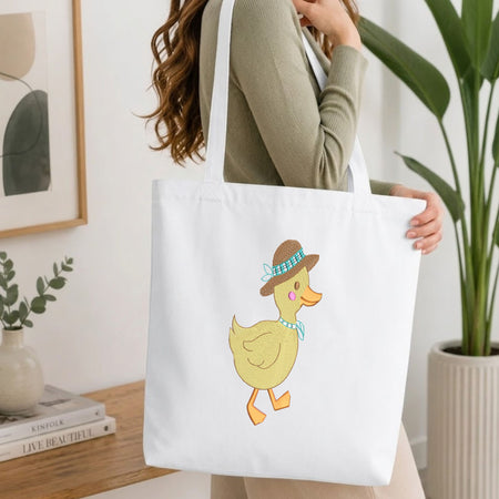 A woman wearing a sage green ribbed top carries a white canvas tote bag featuring a cute yellow duck illustration wearing a brown hat and blue bow tie, set against a bright, modern indoor background with a wooden table and potted plants.